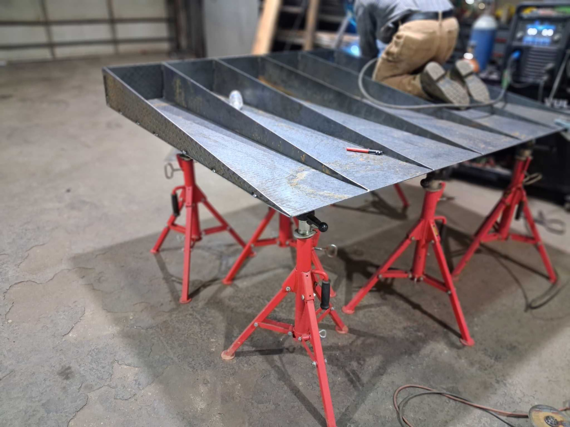 Sheet metal tray with welded divider walls set up on red pipe stands while a welder works behind it