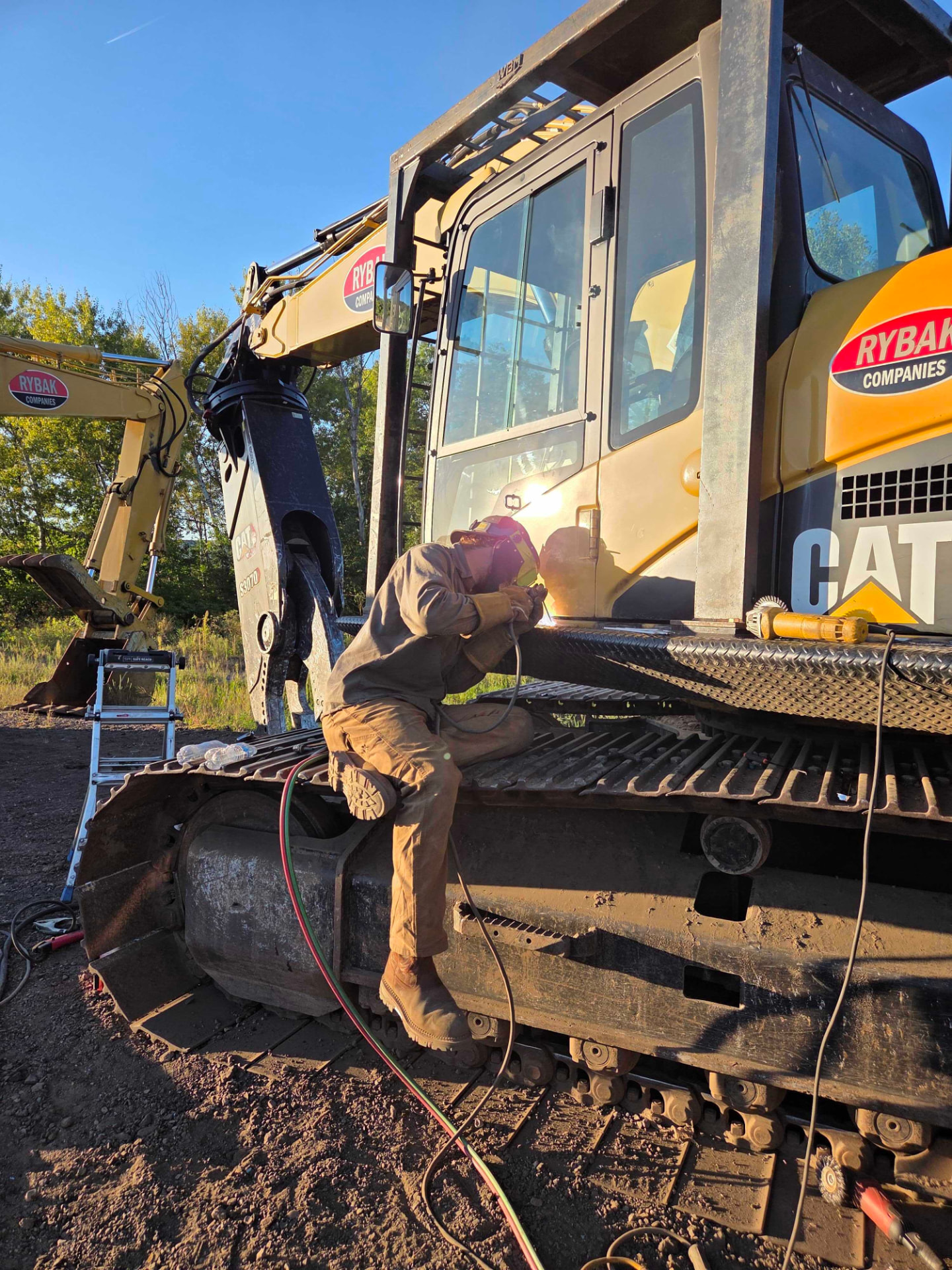 Welder repairing a CAT 336E excavator boom while seated on the track assembly in the field