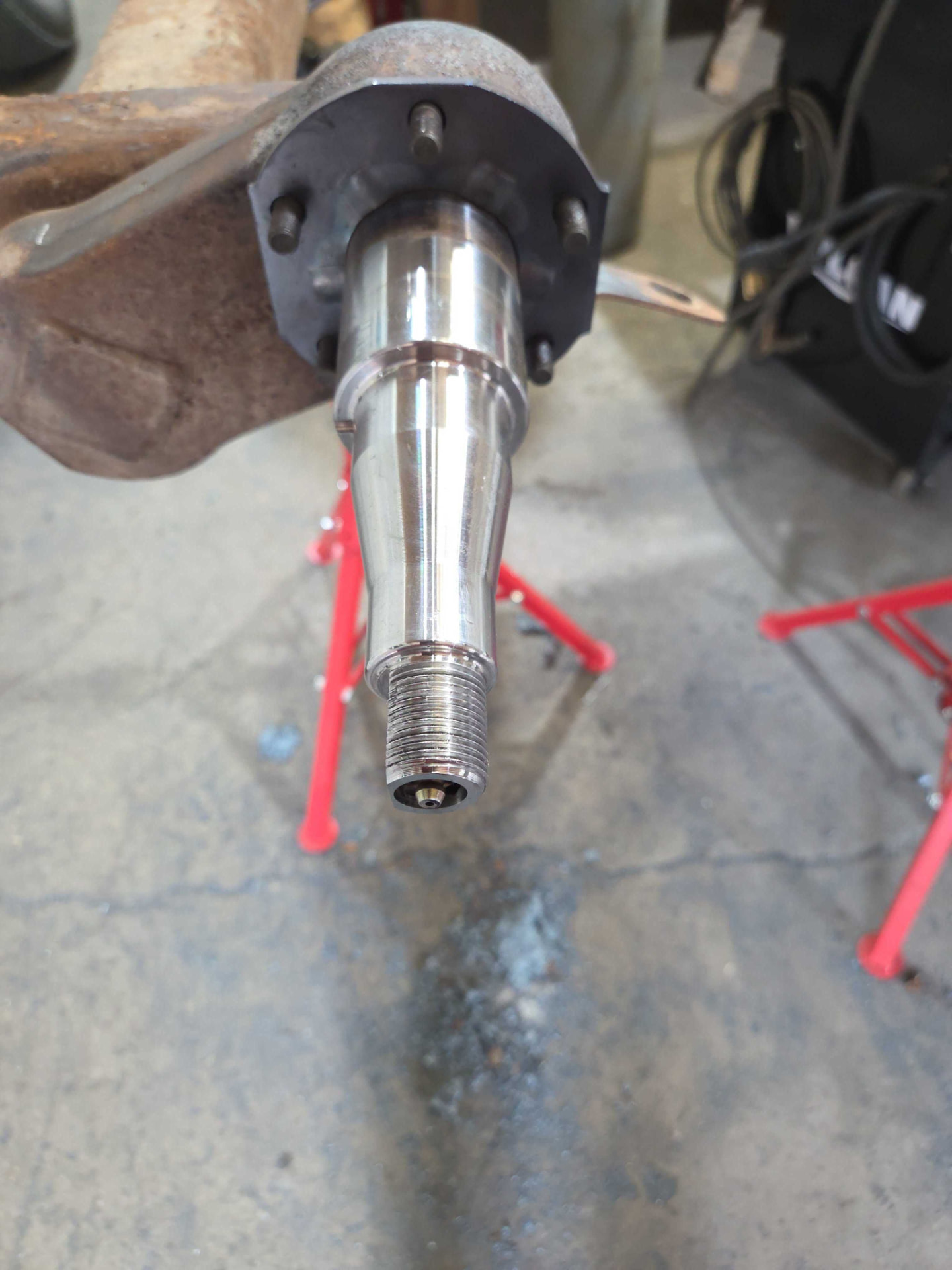 Machined trailer axle spindle with mounting flange and studs, supported on red pipe stands in a shop